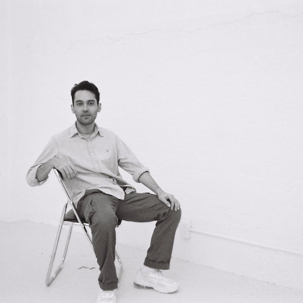A black and white image of Robbie leaning back in a chair in an empty room. Looking at the camera, holding a slight smile. Robbie wears a button up long sleeve top, with the top button undone and sleeves rolled up. He is wearing dark cargo pants with white Nike sneakers. His hand is placed on his knee.